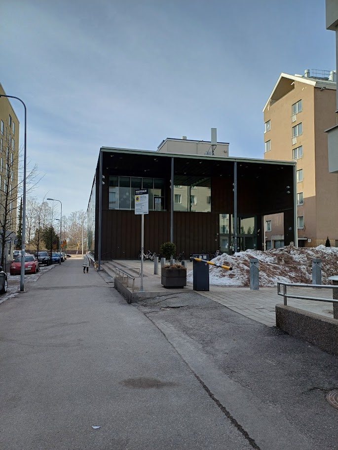 Domus Gaudiumin pääty Leppäsuonkadulta lähestyttäessä: tumma rakennus, jonka päädyssä on leveä katos. Etualalla loiva luiska nousee rakennuksen edustan tasanteelle, ja vasemmalla on kaiteelliset portaat.