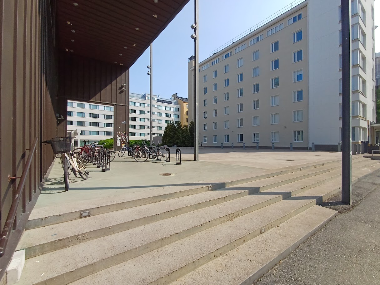 Rakennuksen kulmalla olevat leveät betoniportaat, joiden vasemmassa reunassa seinän vieressä on kaide. Portaiden yläpuolella katoksen alla on tasanne, jolla on pyörätelineitä.
