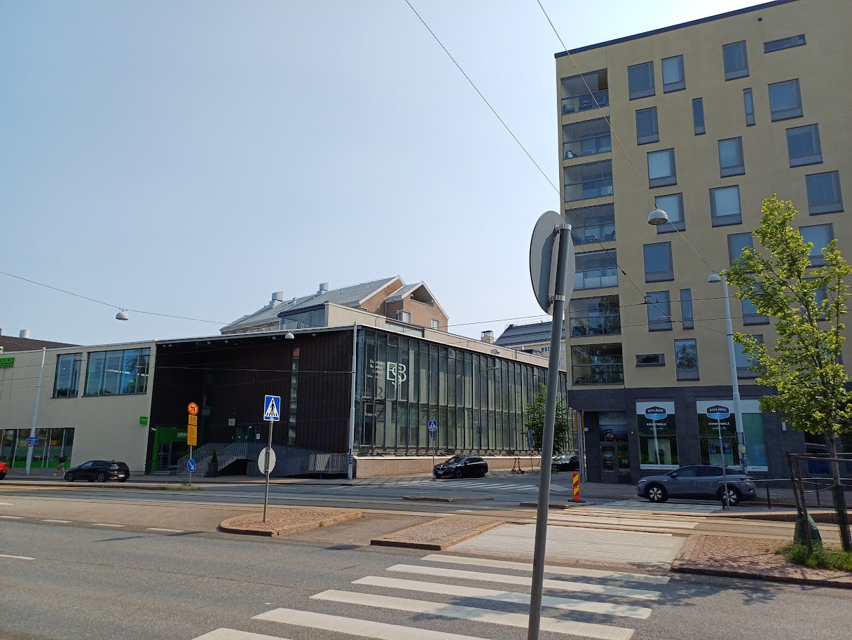 Domus Gaudium Mechelininkadun toiselta puolelta: matala tumma rakennus, jonka kulman lasiseinässä on valkoinen ESB-logo ja vasemmassa päässä vihreä Liikku-kyltti. Oikealla Leppäsuonkadun toisessa kulmassa on vaalea kerrostalo, jonka alakerrassa on Kivilähde-myymälä.