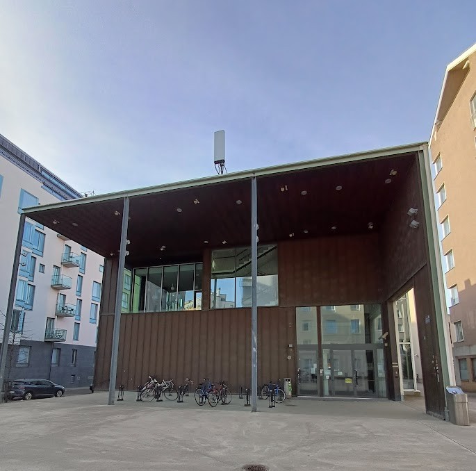 Domus Gaudiumin pääty vastapäätä kuvattuna: ruskea seinä leveän katoksen alla, seinän oikeassa reunassa isot lasiovet. Edustalla on pyörätelineitä.